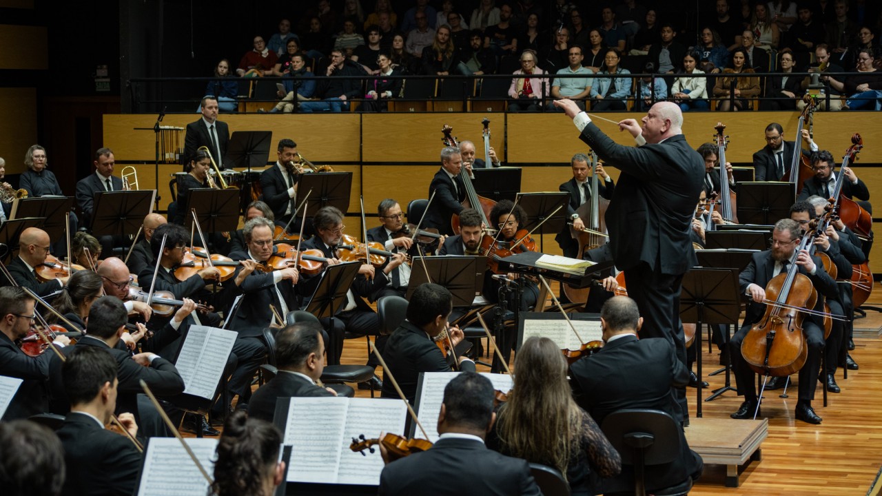 Vemos a orquestra sendo regida por Manfredo Schmiedt. Vemos grande parte da orquestra à esquerda e Manfredo de pé à direita.