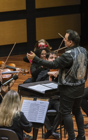 Vemos, &agrave; direita, o maestro Arthur Barbosa com os bra&ccedil;os abertos e sua batuta, regendo a orquestra. &Agrave; esquerda &eacute; poss&iacute;vel ver parte dos jovens da orquestra, a maioria com violinos.