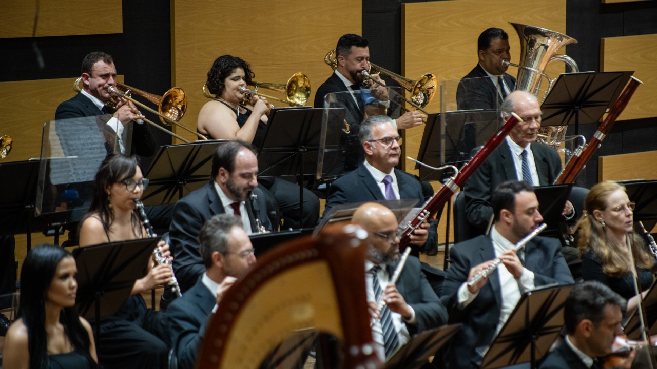 Uma orquestra sinfônica está em apresentação em uma sala de concertos. Ao fundo, há painéis de madeira clara com um padrão retangular. Os músicos estão organizados em fileiras. Na parte superior, estão os instrumentos de sopro de metal, incluindo trombones e uma tuba. Na parte inferior, aparecem músicos com instrumentos de sopro de madeira, como fagotes, flautas e clarinetes. Um pedaço de uma harpa aparece desfocado no canto inferior direito da imagem. Todos os músicos estão vestidos de forma formal, com trajes escuros, e a expressão deles é concentrada, enquanto tocam seus instrumentos.