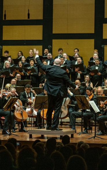 A imagem mostra a Orquestra Sinf&ocirc;nica de Porto Alegre (OSPA) acompanhada de um coro em uma apresenta&ccedil;&atilde;o ao vivo, sob a reg&ecirc;ncia de um maestro. A cena ocorre em um audit&oacute;rio, com a orquestra posicionada na frente e o coro ao fundo. O p&uacute;blico pode ser visto na parte inferior, assistindo &agrave; performance. A composi&ccedil;&atilde;o transmite um momento de intensidade musical, com os m&uacute;sicos e coristas completamente imersos em sua execu&ccedil;&atilde;o.