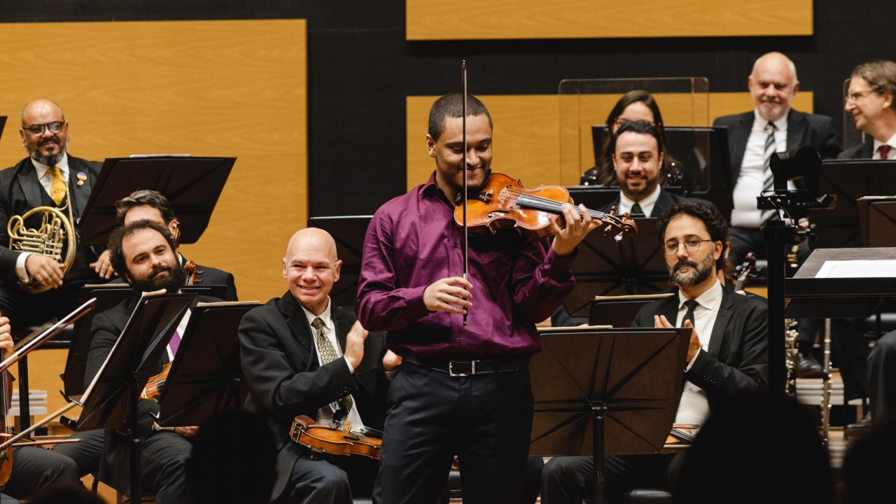 Um violinista está em pé no centro de um palco, segurando um violino de madeira com acabamento brilhante e sorrindo. Ele está vestindo uma camisa de cetim roxa e calça preta. Atrás dele, há uma orquestra composta por músicos vestidos com trajes formais — ternos pretos, camisas brancas e gravatas — segurando instrumentos como violinos, trompas e outros instrumentos de corda e sopro. Alguns músicos estão sorrindo e aplaudindo. O cenário de fundo é composto por painéis de madeira em tons de amarelo e preto, criando um contraste elegante. A atmosfera é de celebração e apreciação musical.