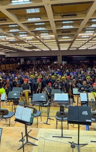 Fotografia em plano aberto tirada do palco da Sala Sinfônica da Casa da OSPA em direção à plateia. A OSPA Jovem posa para uma foto em grupo; os músicos seguram seus instrumentos e muitos usam chapéus coloridos (azul, amarelo, vermelho e verde). Ao fundo, a sala de concertos está lotada com um público que acena para a câmera.
