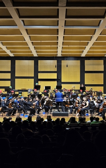 Fotografia panorâmica tirada do fundo da Sala Sinfônica. A orquestra está completa no palco iluminado, disposta em semicírculo sob os painéis acústicos geométricos. A silhueta da plateia sentada ocupa o primeiro plano escuro.