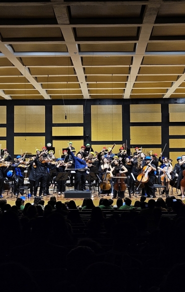 Vista geral do palco a partir da plateia no momento dos aplausos. Os músicos estão de pé com seus instrumentos e arcos levantados para o alto em sinal de celebração. O maestro Arthur Barbosa, ao centro, também está com os braços erguidos.
