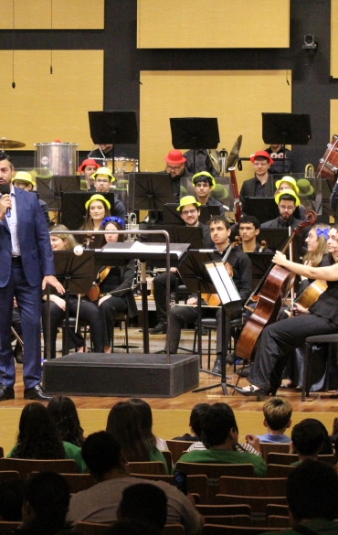 O Diretor da Escola de Música da OSPA, Rodrigo da Rocha, de terno azul fala ao microfone no centro do palco. Atrás dele, os músicos da orquestra estão sentados em suas estantes, usando chapéus coloridos. O fundo é composto por grandes painéis acústicos retangulares em tons de madeira clara e preto.