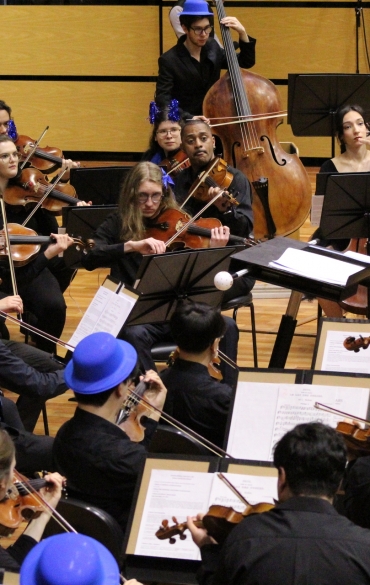 Close na seção de cordas (violinos e violas) da OSPA Jovem durante a execução musical. Os músicos, vestidos de preto e usando chapéus de coco azuis, tocam seus instrumentos sob a regência do maestro Arthur Barbosa, de terno azul, que é visto de costas à direita da imagem.