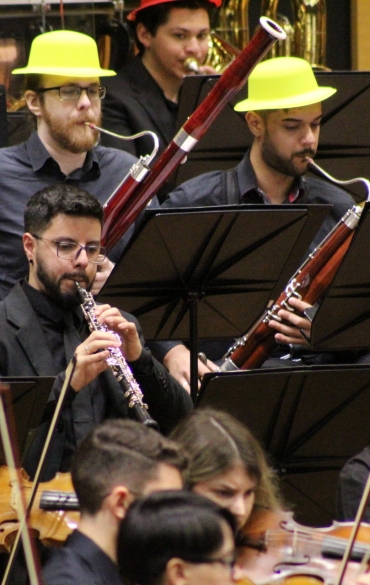 Plano médio focado na seção de sopros de madeira. No centro, um músico de barba e óculos toca oboé, cercado por colegas que tocam fagote. Todos os músicos visíveis nesta seção usam chapéus amarelos vibrantes.