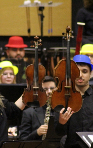 Dois músicos, um homem e uma mulher usando chapéus azuis, estão de pé no palco segurando um violino e uma viola lado a lado. A imagem destaca a diferença de tamanho entre os dois instrumentos de cordas para fins didáticos.