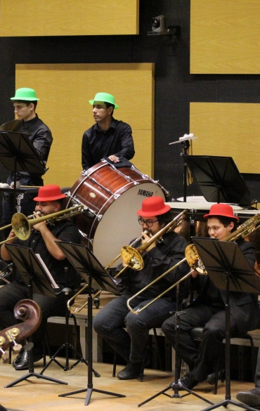 Vista lateral da seção de metais e percussão da OSPA Jovem. Na frente, músicos tocam trombones e usam chapéus vermelhos. Ao fundo, em um nível elevado, os percussionistas usam chapéus verdes. À direita, destacam-se grandes tímpanos de cobre polido.