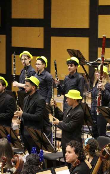 Músicos da seção de sopros (flautas, clarinetes, oboés e fagotes) em pé no palco, segurando seus instrumentos verticalmente em uma pose de apresentação. Eles vestem preto e usam chapéus amarelos.