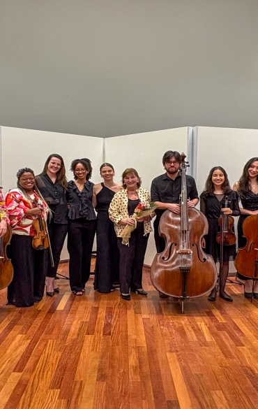 Doze alunos da Escola da OSPA posam em pé com seus instrumentos junto à professora Olinda Alessandrini na Sala de Recitais da Casa da OSPA, com piso de madeira e painéis acústicos brancos ao fundo. Eles seguram instrumentos de cordas, como violinos, violas, violoncelos e um contrabaixo acústico. Olinda segura um buquê de flores.