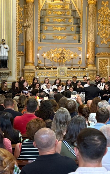Vista de trás de uma plateia sentada em uma igreja ricamente decorada com detalhes em dourado e esculturas sacras. Ao fundo, no altar, o Coro Jovem e Coro Infantojuvenil da Escola de Música da OSPA se apresentam sob a regência do maestro Cosmas Grieneisen, de costas para o público.