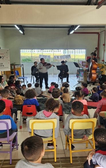 Um grupo de crianças pequenas sentadas em cadeiras coloridas em uma sala de aula de educação infantil assiste a uma apresentação musical. À frente, músicos da Escola da OSPA tocam violino, violoncelo e contrabaixo. Há um banner do projeto "Escola da OSPA na Comunidade".