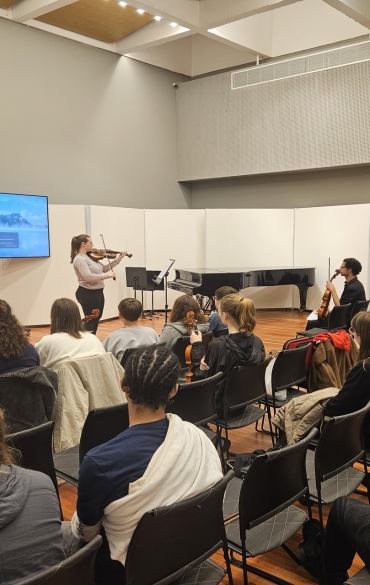 Uma violinista se apresenta em pé em na Sala de Recitais da Casa da OSPA para uma plateia de estudantes sentados. À esquerda, uma tela de TV exibe uma imagem de paisagem. Ao fundo, um piano de cauda preto.