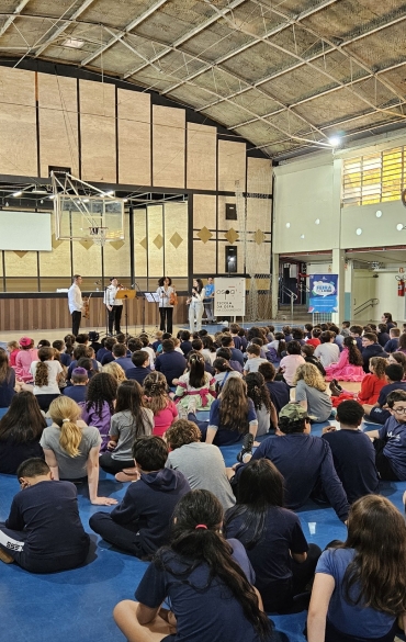Uma ampla plateia de crianças sentada no chão de um ginásio poliesportivo azul assiste a quatro músicos da Escola de Música da OSPA que se apresentam ao fundo. O ambiente é iluminado e possui estruturas de basquete.