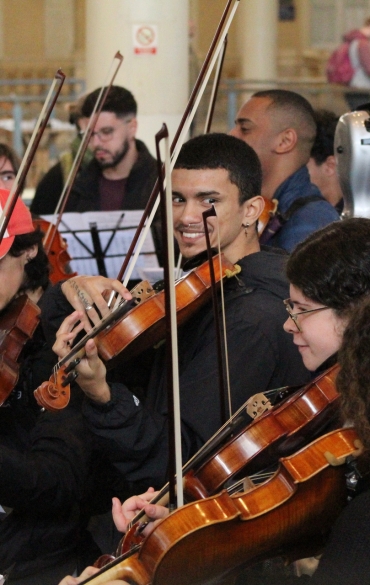 Close-up de músicos da OSPA Jovem tocando violino no Mercado Público de Porto Alegre. Um jovem violinista no centro sorri para a câmera enquanto toca. Pessoas ao redor registram o momento com celulares e câmeras profissionais.