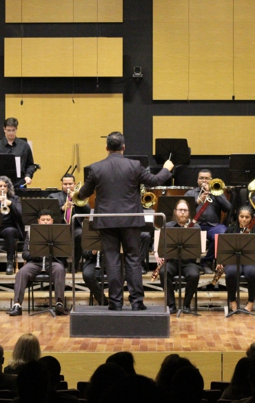 Visão frontal da Orquestra de Sopros da Escola da OSPA em um palco. O maestro Wilthon matos, de costas para a câmera, rege o grupo composto por instrumentos de metal, madeira e percussão. O fundo do palco possui painéis retangulares amarelos e pretos.