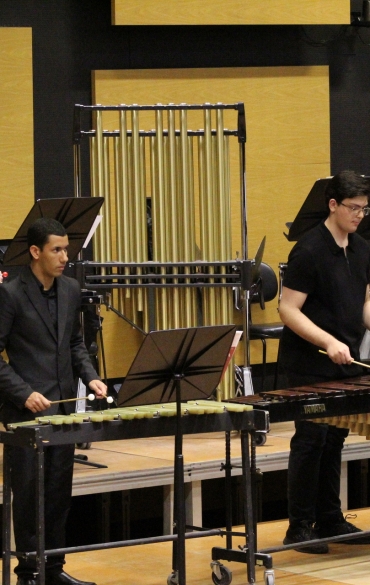 Quatro percussionistas da Orquestra de Sopros em ação no palco. Um músico toca carrilhão (sinos tubulares), outro toca xilofone, o terceiro toca marimba e o quarto está ao lado de tambores. Eles vestem roupas pretas formais.