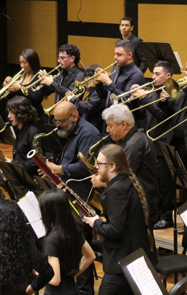 Close lateral de uma seção da Orquestra de Sopros. Músicos jovens tocam simultaneamente fagotes, saxofones, trompetes, trombones e tubas. O foco está na concentração dos instrumentistas durante a execução.