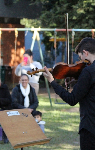Em um ambiente externo com gramado e um parquinho infantil ao fundo, dois jovens músicos da Escola de Música da OSPA, vestidos de preto, tocam violino de pé. Eles estão posicionados de perfil e de costas para a câmera, voltados para uma plateia composta por crianças e adultos que assistem à apresentação sentados na grama e em brinquedos. Uma estante de partitura de madeira está posicionada à frente dos músicos.