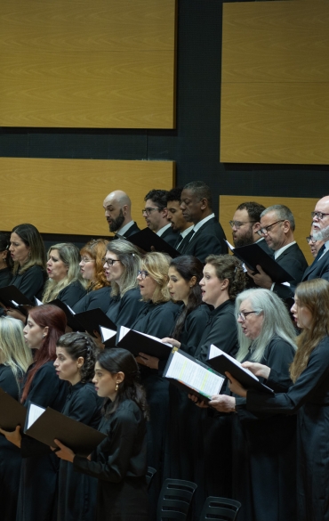 Um coro de trinta integrantes, vestindo trajes formais pretos, canta em um palco. O grupo est&aacute; organizado em fileiras, segura pastas de partitura e demonstra concentra&ccedil;&atilde;o. Ao fundo, a parede escura apresenta grandes pain&eacute;is ac&uacute;sticos de madeira clara dispostos de forma retangular.