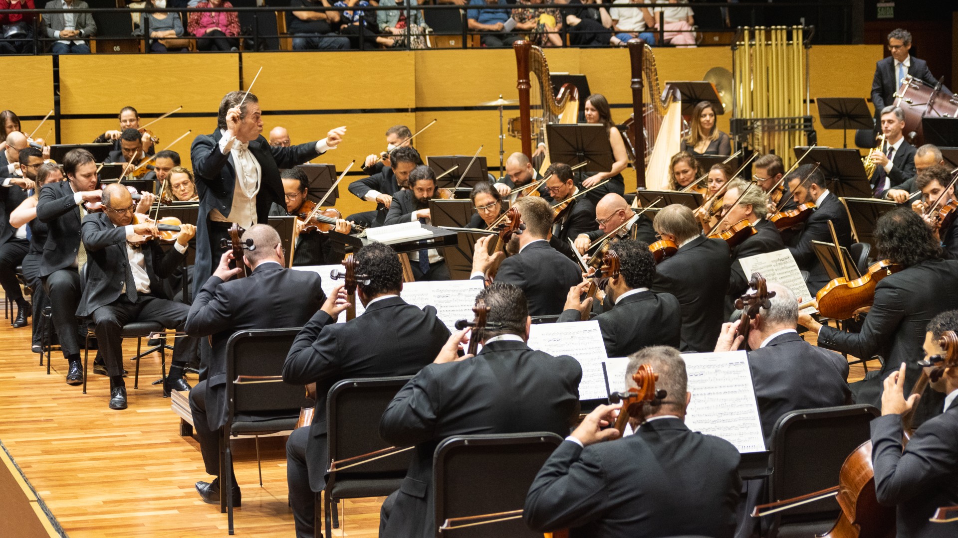 A fotografia registra um concerto da Orquestra Sinf&ocirc;nica de Porto Alegre (OSPA), com o maestro Jos&eacute; Mar&iacute;a Moreno em destaque no lado esquerdo, regendo com gestos expressivos e uma batuta. No palco de madeira clara, dezenas de m&uacute;sicos em trajes formais tocam diversos instrumentos, com foco principal nos violinos e violoncelos em primeiro plano. Ao fundo, v&ecirc;-se o coro e a plateia acompanhando a performance em uma sala de concertos iluminada e moderna.