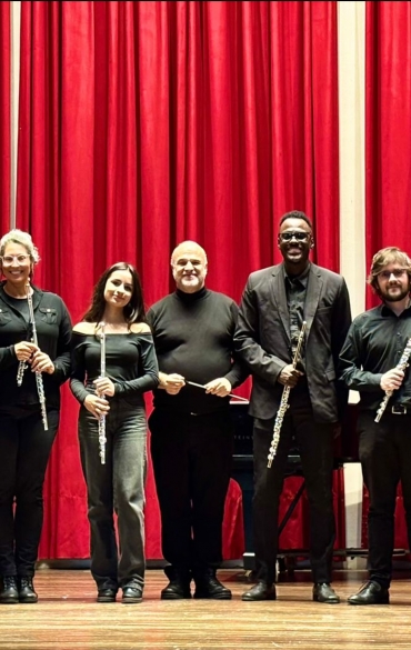 Um grupo de nove m&uacute;sicos posando alinhados no palco, em frente a uma grande cortina vermelha de teatro. H&aacute; oito flautistas (segurando flautas) e um maestro ao centro (segurando uma batuta). Todos est&atilde;o vestidos com roupas formais em tons escuros (preto e cinza-escuro). O ch&atilde;o &eacute; de madeira clara e o fundo &eacute; composto inteiramente pela cortina vermelha plissada. Todos sorriem para a foto.