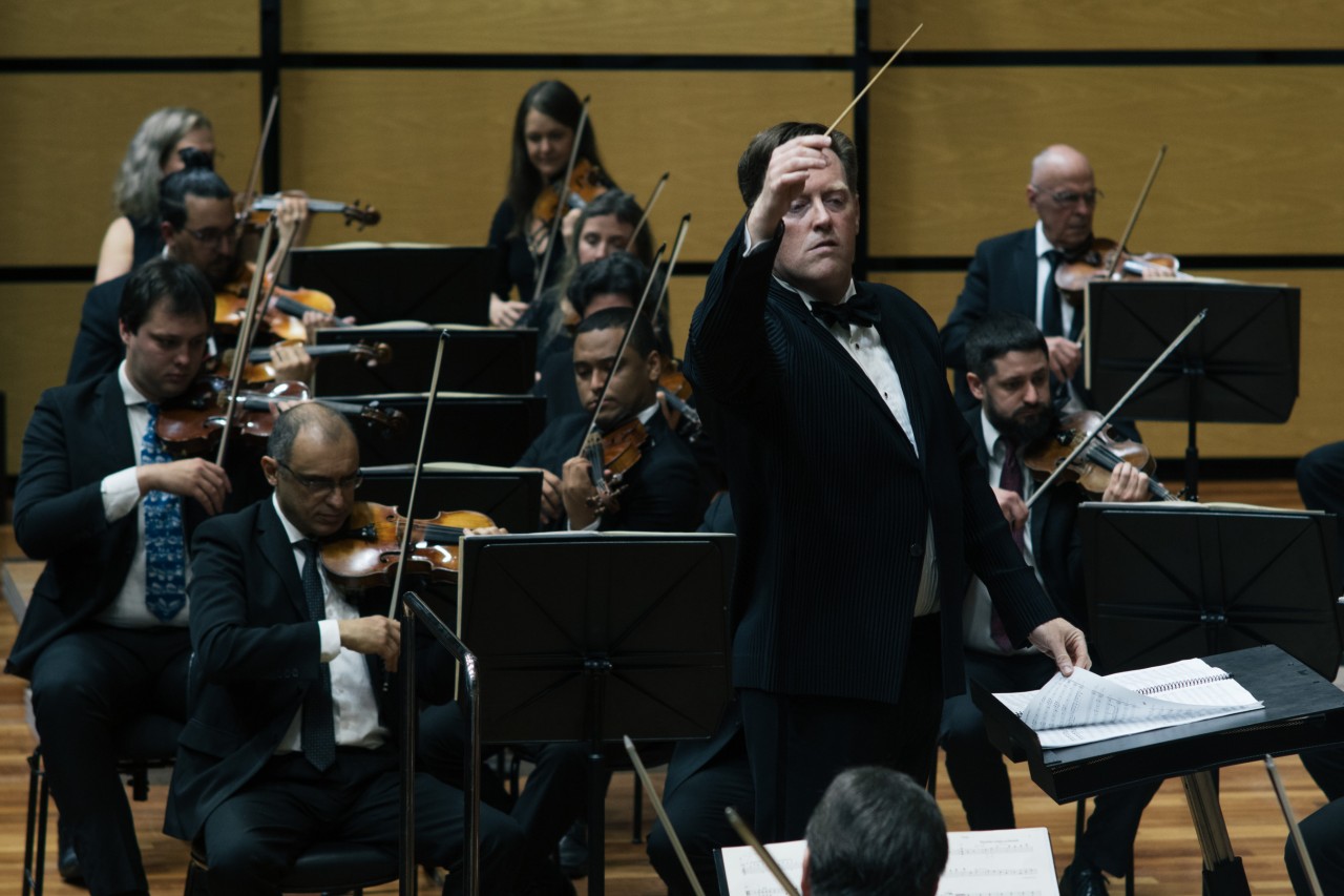 Maestro Jonathan Girard ergue a batuta diante de alguns músicos da OSPA, que tocam violinos ao fundo, na Sala Sinfônica da Casa da OSPA.