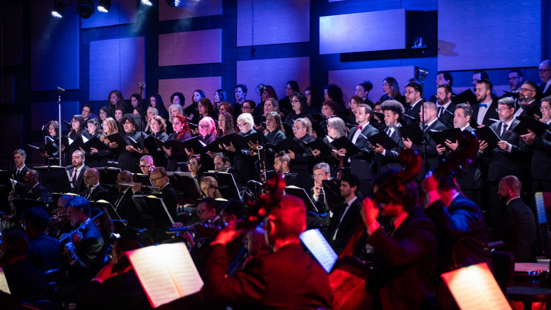 Sob luz azul, o Coro Sinf&ocirc;nico da OSPA est&aacute; em p&eacute; no fundo da Sala Sinf&ocirc;nica. Em frente, aparecem v&aacute;rios m&uacute;sicos sentados tocando instrumentos variados. Cena do espet&aacute;culo Cl&aacute;ssicos da Broadway.